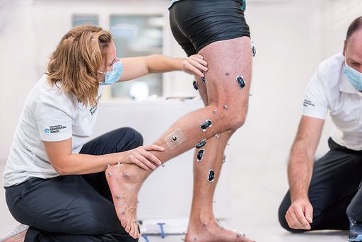 In der Hocke eine Medizinerin, die Sensoren an die Beine eines Patienten anbringt.