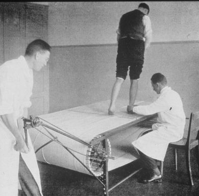 Historisches Bild in schwarz-weiss: Der Patient steht auf einem Laufband, während ein Mediziner den Fuss des Patienten hält und ein anderer Mediziner die Kurbel des Laufbandes betätigt.